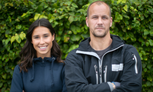 To personer står foran grønn hekk, kledd i mørke jakker