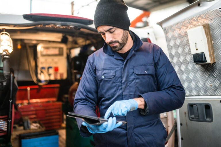 Person i blå kjeledress og hansker bruker nettbrett foran servicebil