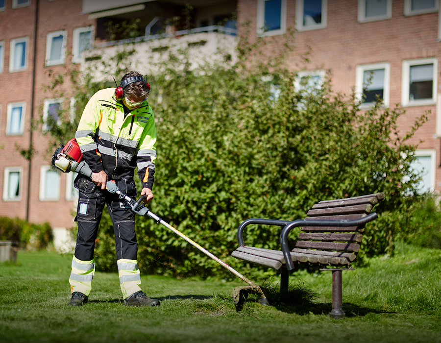Person i verneutstyr trimmer gress rundt benk i park.