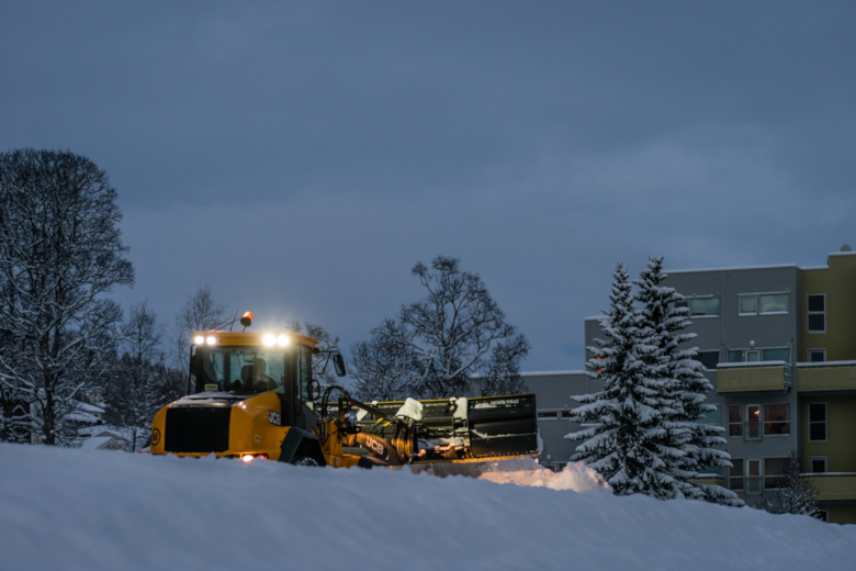 Gul snøplog rydder snø langs vei foran bygninger i vinterlandskap.